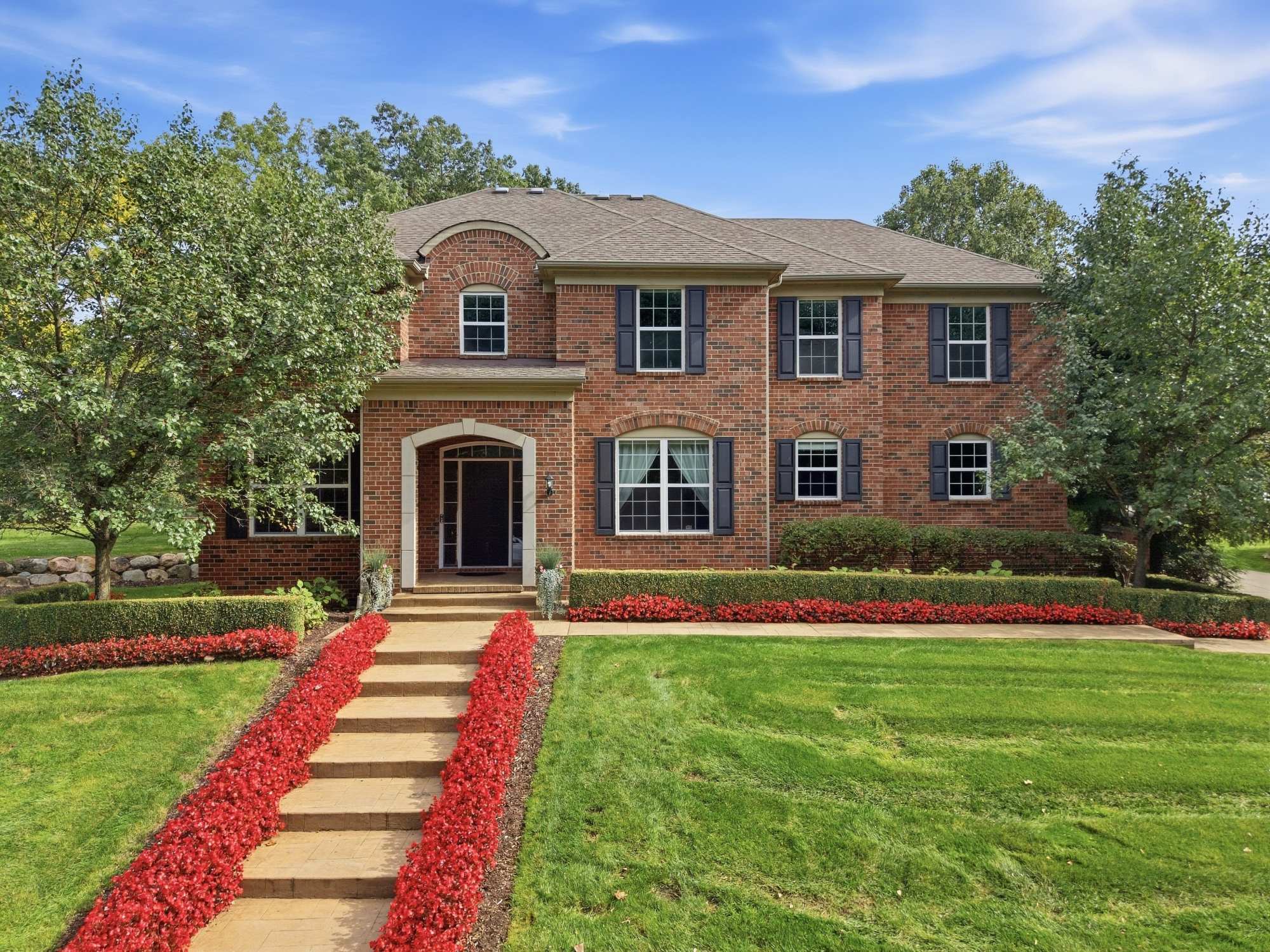 319 Greenan Lane, a red brick colonial with a tiered stone walkway and red begonia beds, seen from an elevated angle under a blue sky